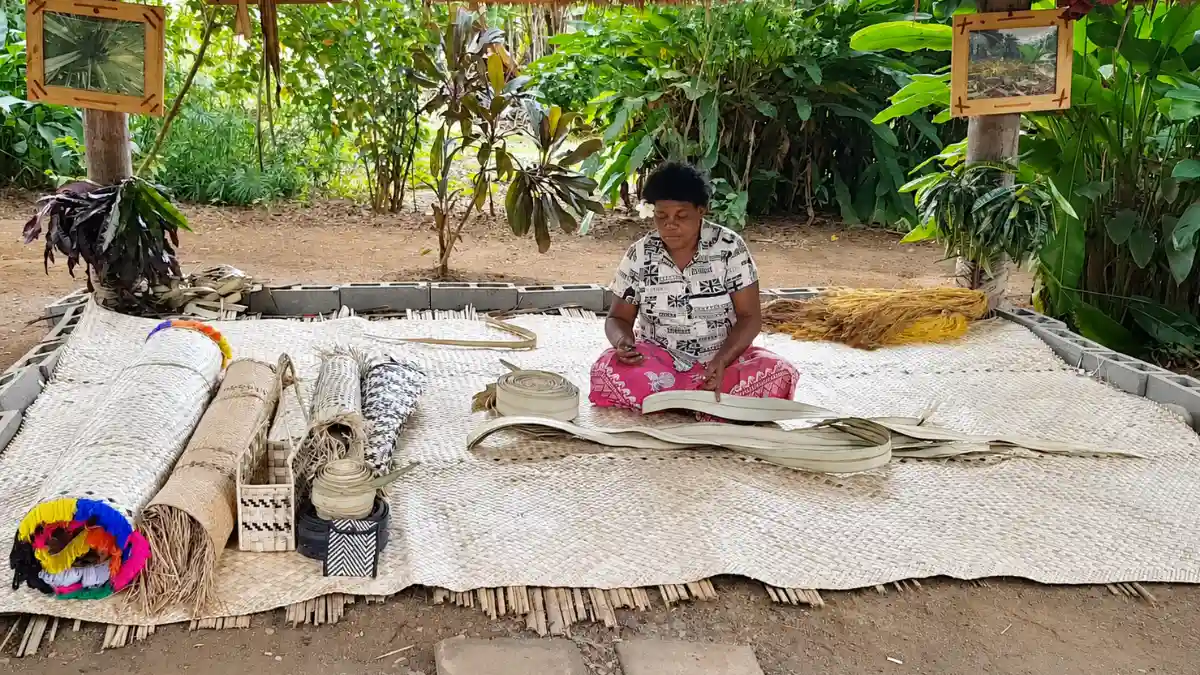 A woman weaving leaf strips beside handwoven mats and baskets