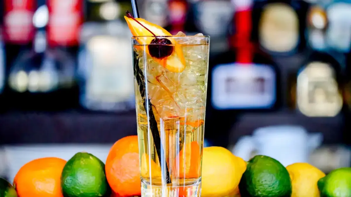 Tall iced cocktail garnished with an orange slice and cherry at a bar