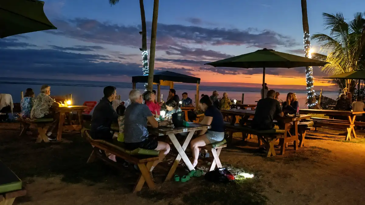 Guests dining at outdoor picnic tables beside the water at sunset at Crab Shack Fiji in Korotogo